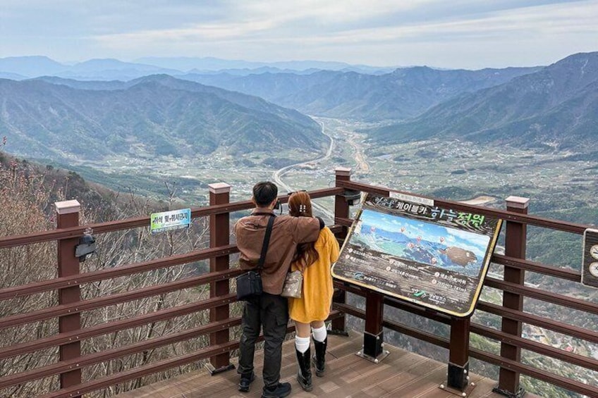 A couple standing at a viewpoint with a signboard, overlooking a valley and mountains. 3