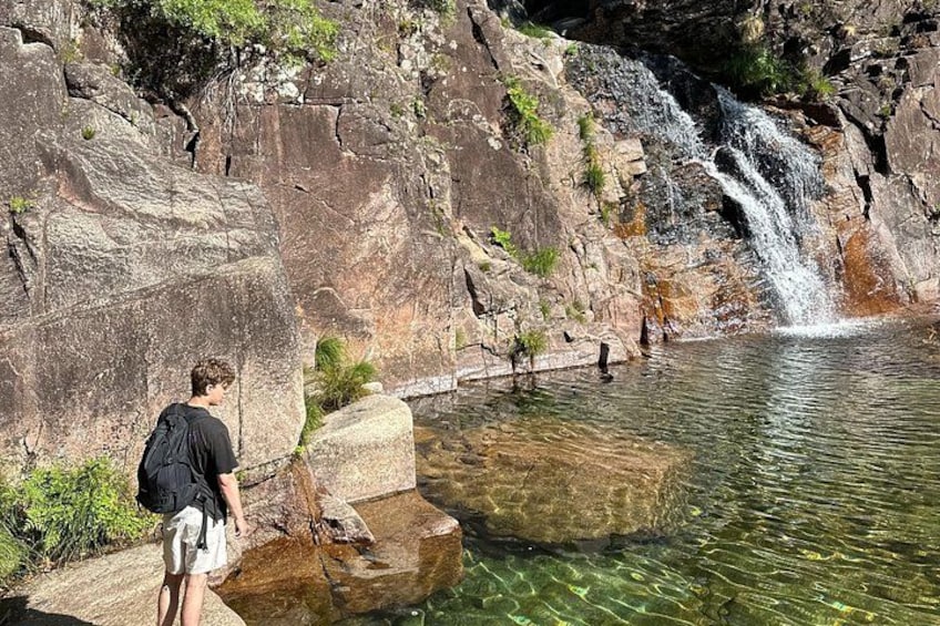A waterfall, clear water, rocky terrain, and a person standing by the water. 1