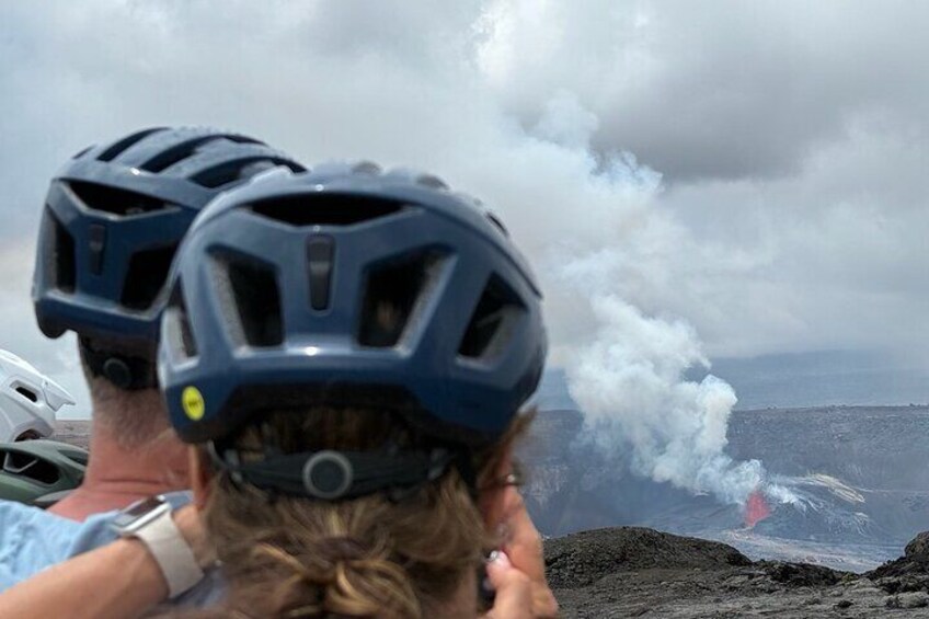 Two people wearing helmets observing a volcanic eruption with smoke and lava visible. 1