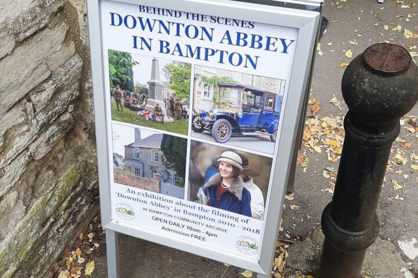 A signboard advertising the 'Downton Abbey in Bampton' exhibition with photos of the film's locations and a historical building. 4