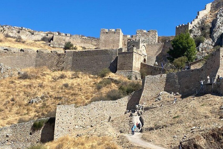 A historical stone fortress with walls and towers, a dry grassy landscape, and a clear blue sky. 5