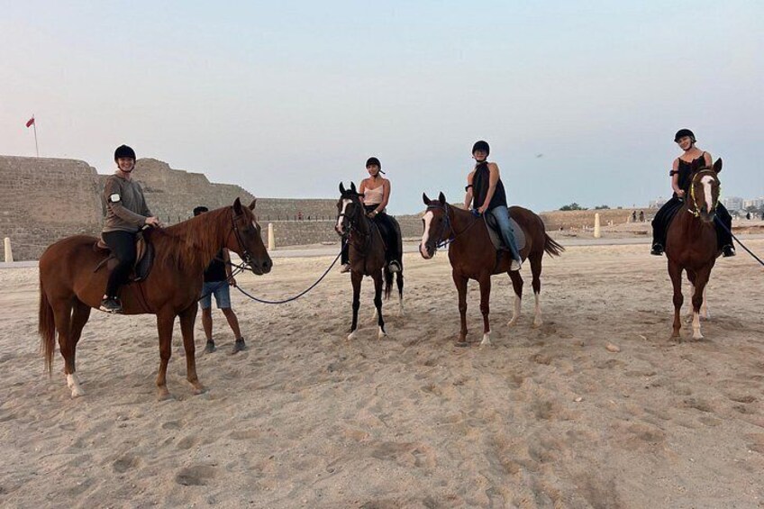 A group of people riding horses on a sandy beach with a large wall in the background. 5