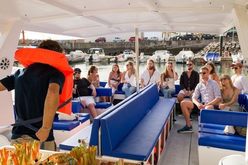 Eine Gruppe von Menschen auf einem Boot mit blauen Sitzen, eine Person trägt eine Rettungsweste, und im Hintergrund liegt eine Marina mit Booten. 2