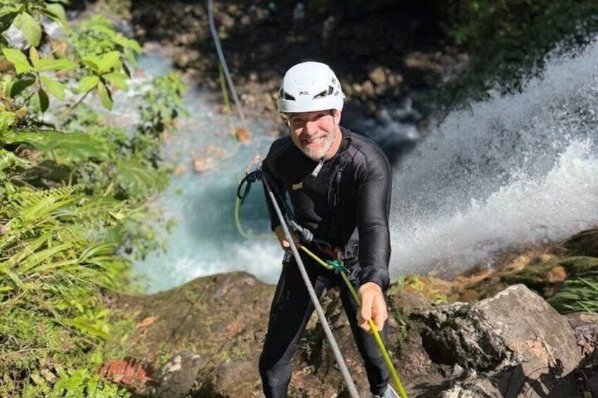 Eine Person seilt sich an einer steilen, felsigen Klippe ab, umgeben von üppiger Vegetation und einem Fluss. 1