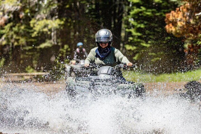 A person riding an ATV through a body of water, splashing water around. 3
