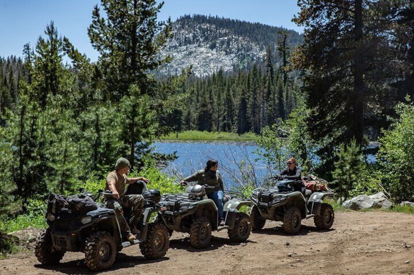 Three off-road vehicles parked on a dirt path near a lake surrounded by trees and mountains. 2