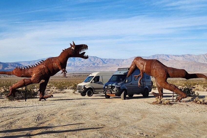 Ein großes Dinosaurier-Modell, ein Fahrzeug und eine Wüstenlandschaft mit Bergen im Hintergrund. 5