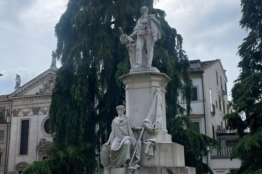 A statue of a standing figure with a seated figure below, surrounded by trees and buildings. 2