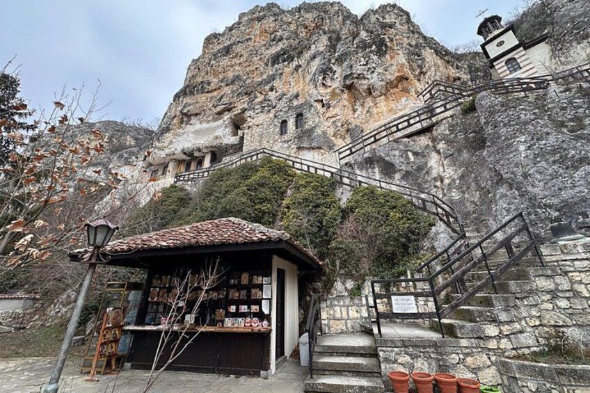 A stone staircase leading up to a building with a tiled roof, surrounded by rocky cliffs and sparse vegetation. 1