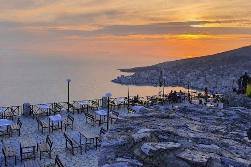 A coastal sunset view with stone tables and benches, rocky terrain, and a distant cityscape. 4