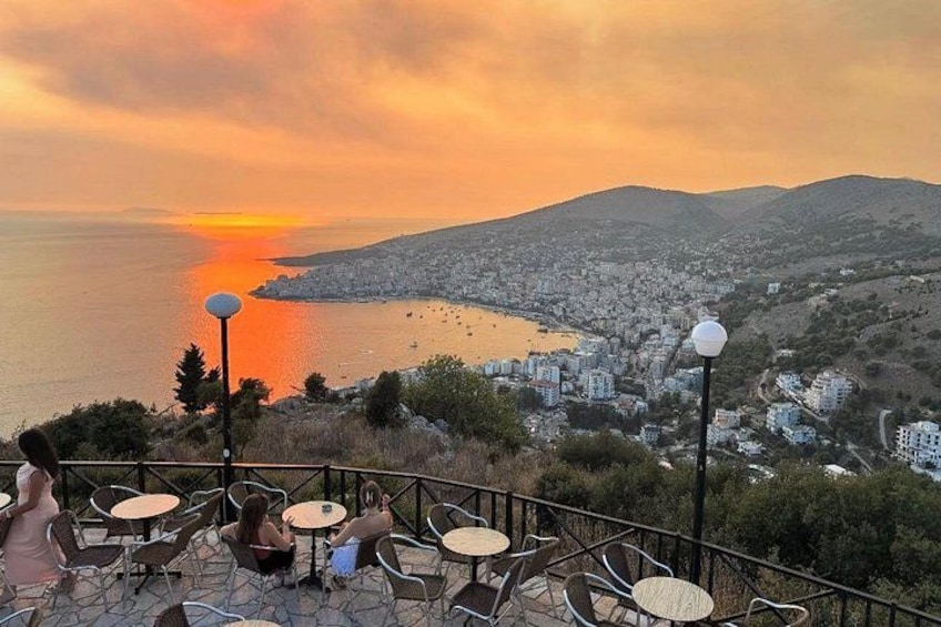 A sunset view over a coastal city with chairs and tables on a terrace. 1