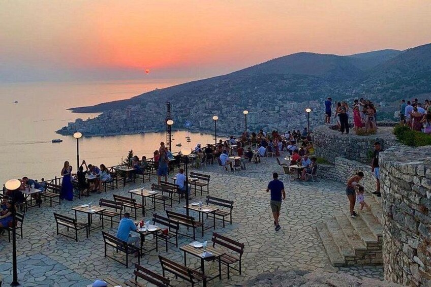 A coastal terrace with tables and chairs, overlooking a city and the sea at sunset. 5