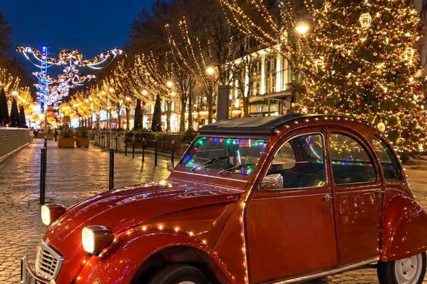 A vintage car parked on a cobblestone street, decorated with Christmas lights, with festive trees and lights in the background. 4