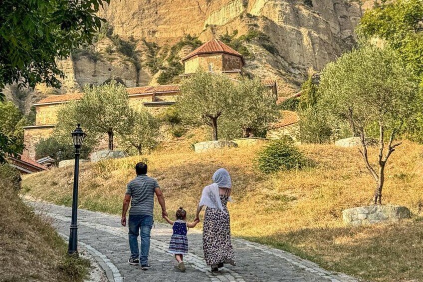 Eine Familie spaziert auf einem Kopfsteinpflasterweg in einer hügeligen Landschaft, im Hintergrund Olivenbäume und Gebäude. 4
