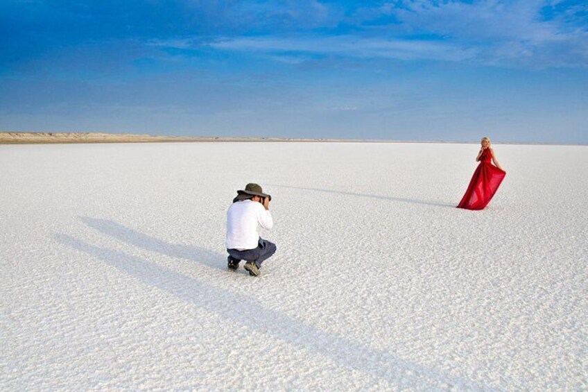 A person in a white shirt and hat crouching, taking a photo of another person in a red dress standing in a vast white landscape. 1