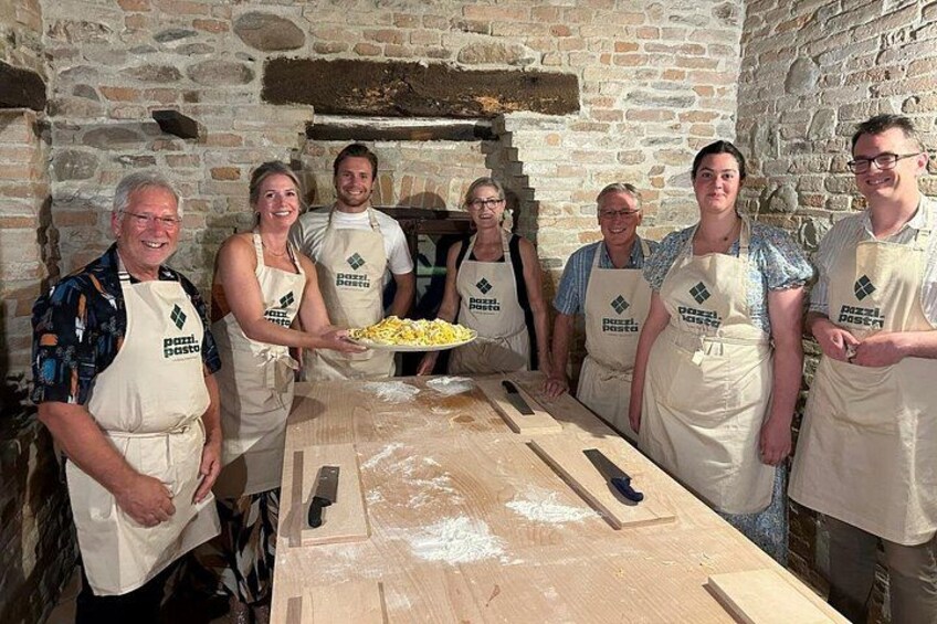 A group of people in aprons in a rustic kitchen with stone walls, a wooden table, and a pizza oven. 5