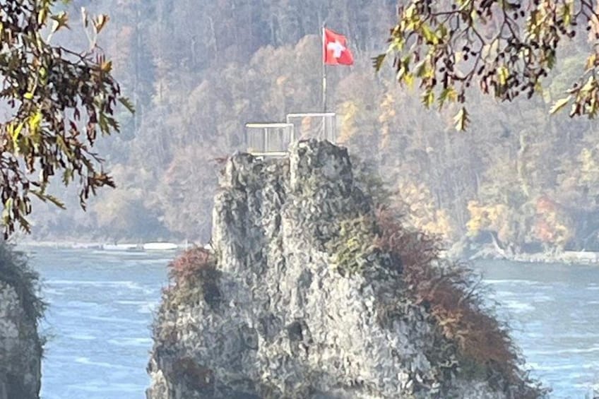 A rocky cliff with a flag, surrounded by water and trees. 1