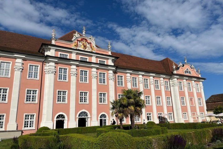 A large pink and white building with columns and a crest, surrounded by well-manicured hedges and palm trees. 4