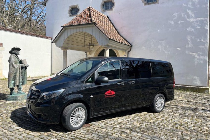 A black minivan parked on cobblestone pavement in front of a white building with a red-tiled roof. 5