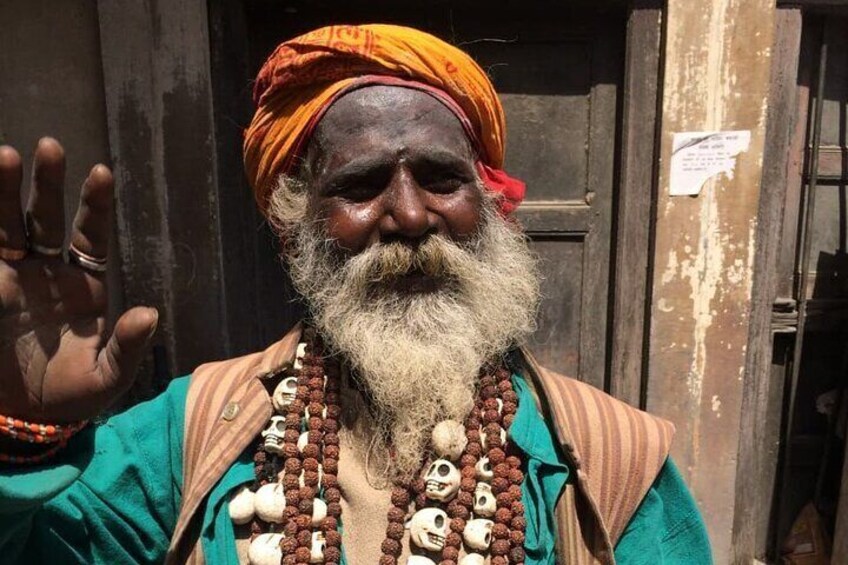 A person with a long white beard, wearing a turban and multiple necklaces with beads and skulls. 1
