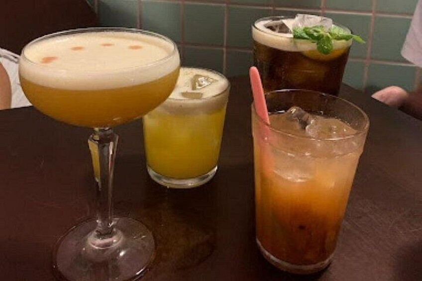 Three drinks on a table: a cocktail in a stemmed glass, a glass with a yellow liquid and a lime wedge, and a tall glass with a dark liquid and a mint garnish. 3