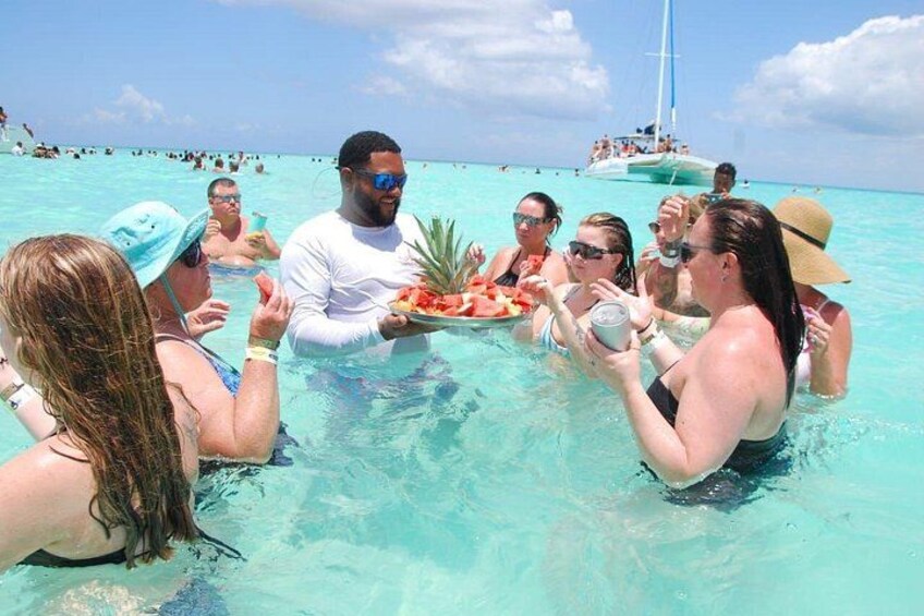 A group of people in a shallow, clear blue sea, some holding plates of food, with a sailboat in the background. 2
