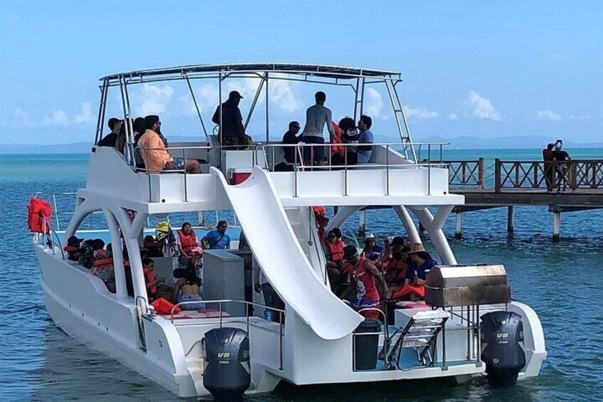 A white boat with a covered upper deck and a slide, filled with people, docked at a pier. 2