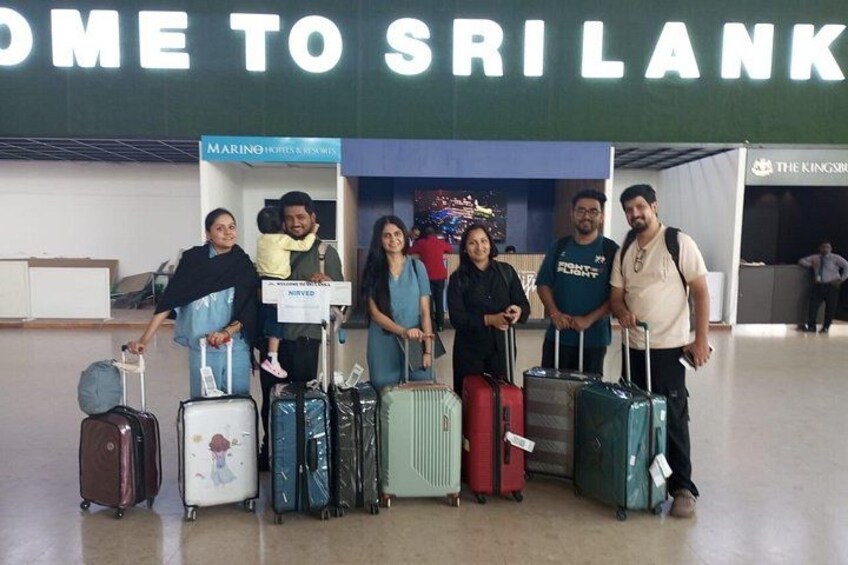 A group of people with luggage at an airport, with a sign reading 'HOME TO SRI LANKA' in the background. 2