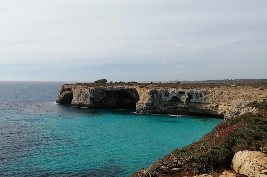 A coastal cliff with a natural arch formation, clear turquoise waters, and sparse vegetation. 5