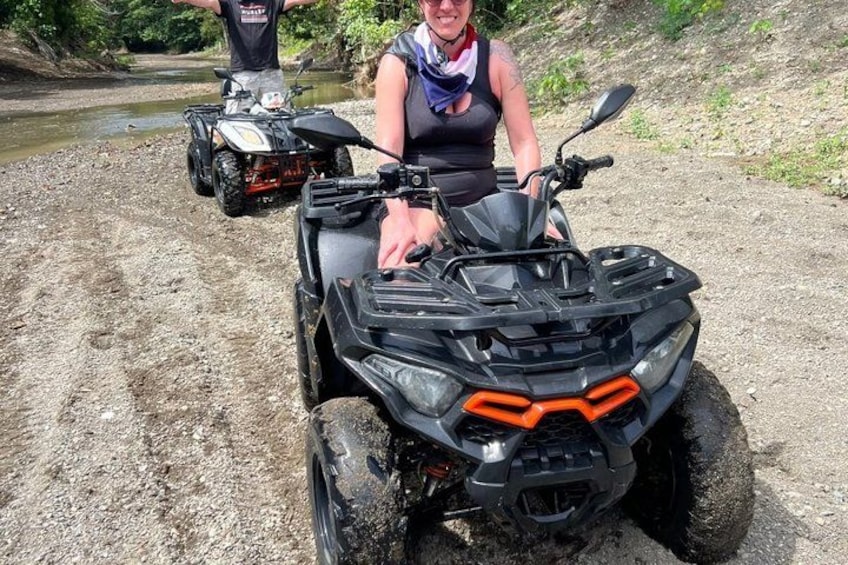 Eine Person fährt einen schwarzen ATV mit orangefarbenen Akzenten, im Hintergrund ein weiteres ATV und ein Fluss. 3