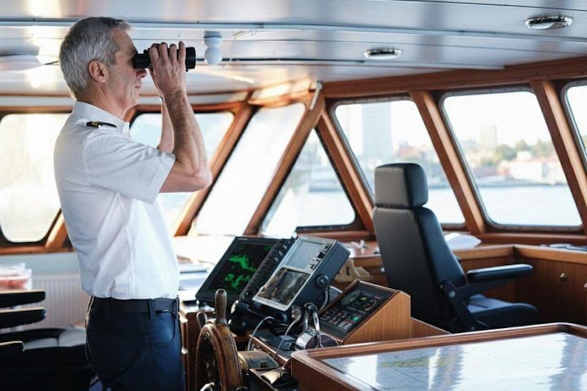 A man using binoculars inside a boat with a control panel and windows. 3