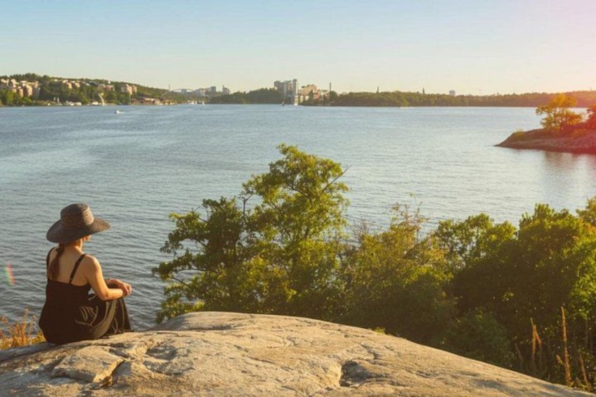 A person sitting on a rock by a calm river with trees in the foreground and a cityscape in the background. 2