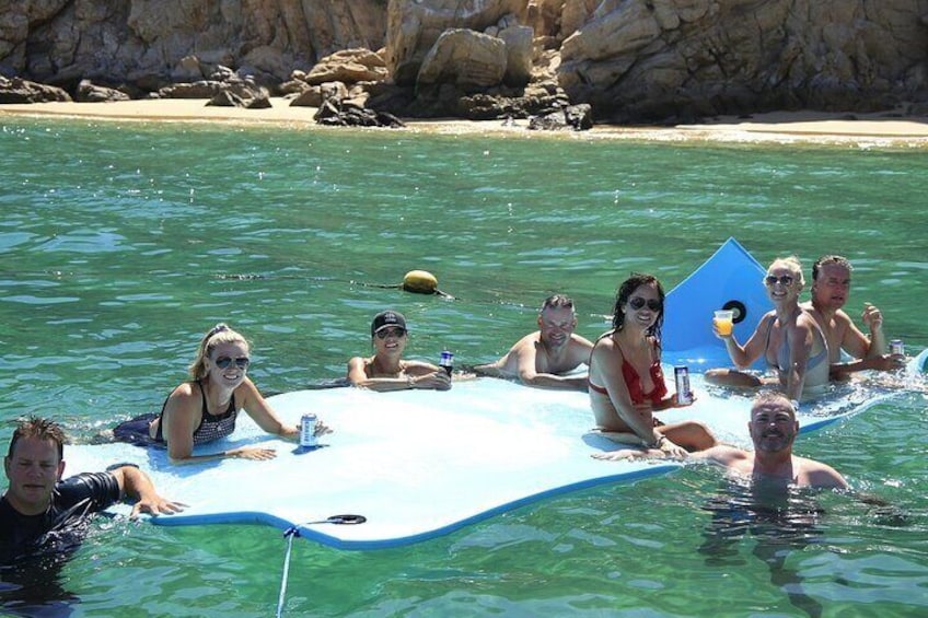 A group of people floating on a blue raft in clear water with a rocky shoreline in the background. 4