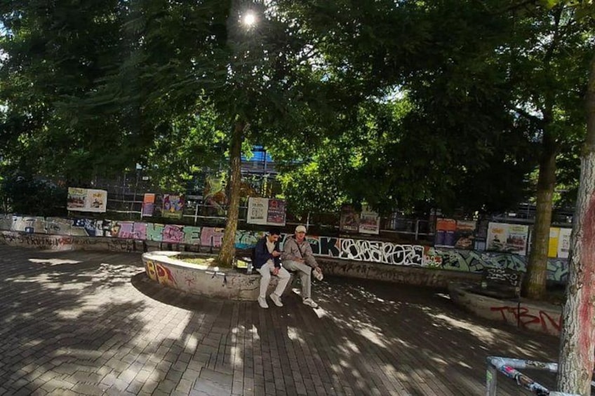 A person sitting on a bench in a park with graffiti-covered walls and trees. 5