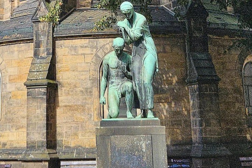 A bronze statue of a man kneeling and praying, with a woman standing beside him, set against a stone building with arched windows. 4