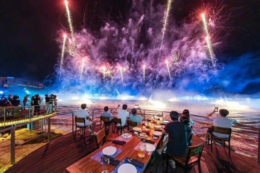 A dining area with tables and chairs, overlooking a body of water with fireworks in the background. 3