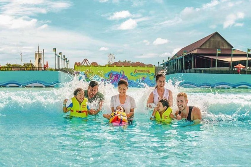 Eine Gruppe von Menschen in einem Wasserpark mit einem Wellenbecken und einem Gebäude im Hintergrund. 5
