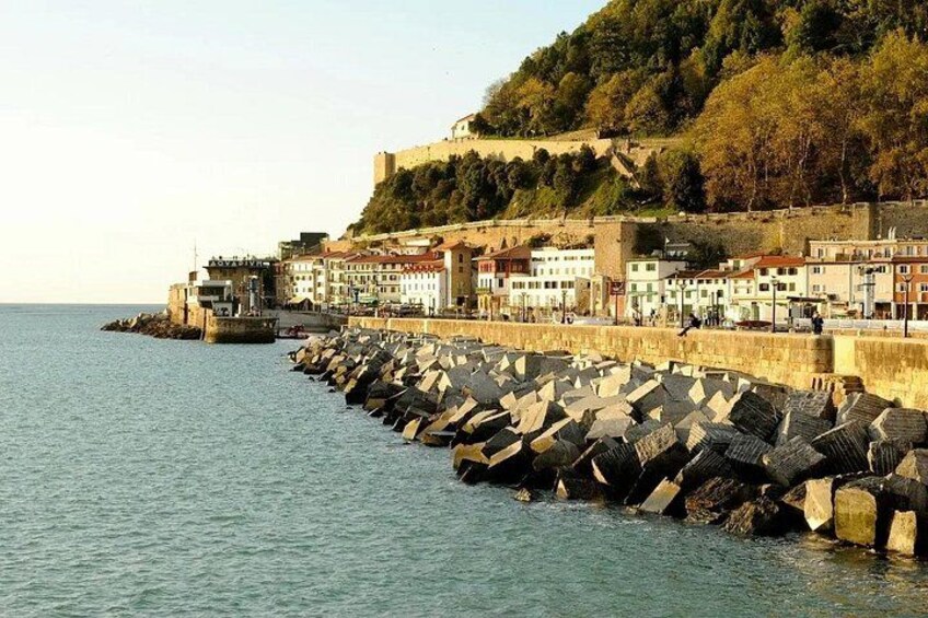 A coastal town with a stone sea wall, buildings, and a hill with trees. 3
