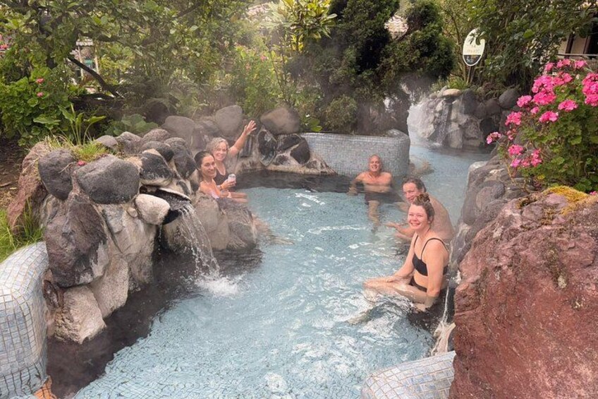 Eine heiße Quelle mit entspannten Menschen im Wasser, umgeben von Felsen und Grünpflanzen. 1