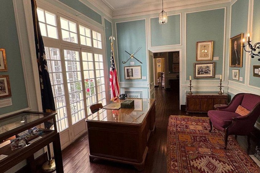 A room with a wooden desk, a red armchair, a patterned rug, and framed pictures on the walls. 2