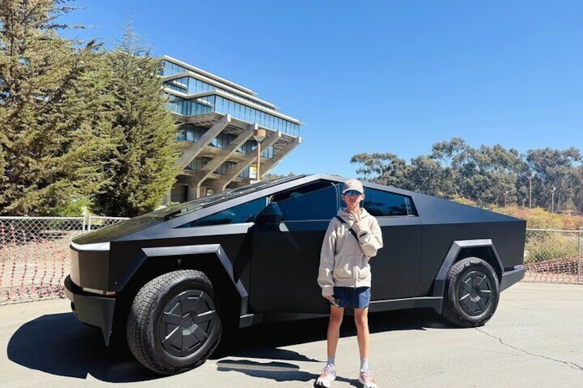 A person standing next to a futuristic vehicle with a unique design, large tires, and a modern building in the background. 2