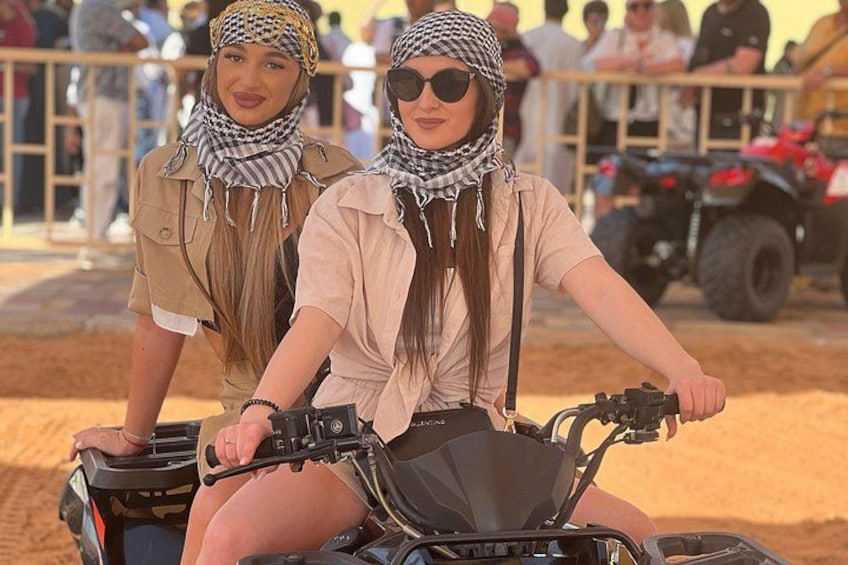 Two people on an ATV in a sandy area with spectators and vehicles in the background. 1