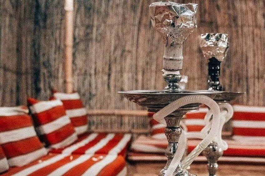 A hookah with a metallic base and a crystal glass on top, set against a wooden wall with striped cushions. 2