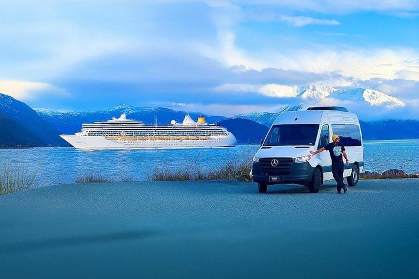 A white van parked by a body of water with a cruise ship in the background and snow-capped mountains. 2