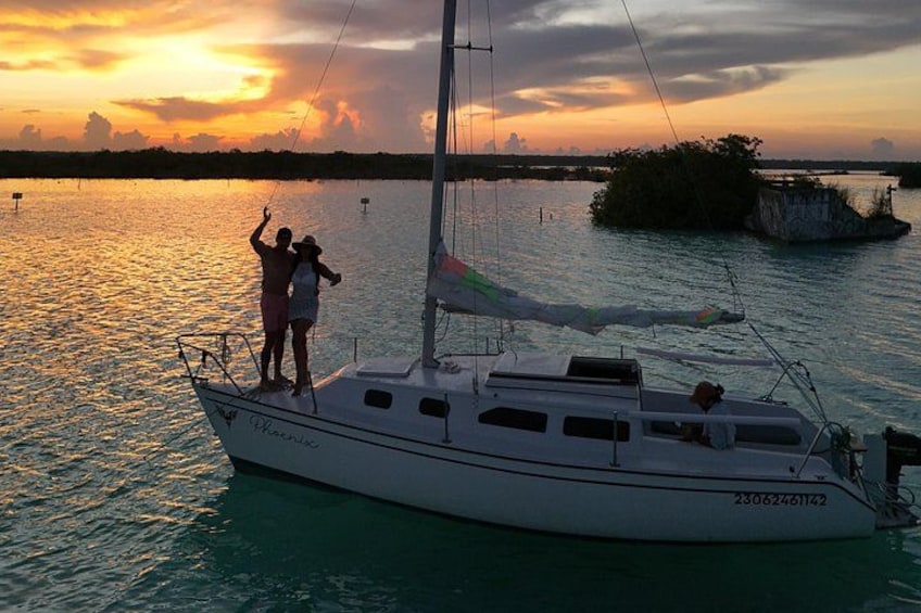 A sailboat with two people standing at the bow, a calm sea, and a sunset in the background. 2