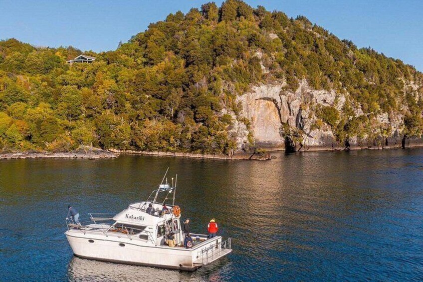 A boat named 'Kaharik' on calm water, a forested hill in the background, and a small house on the hill. 5