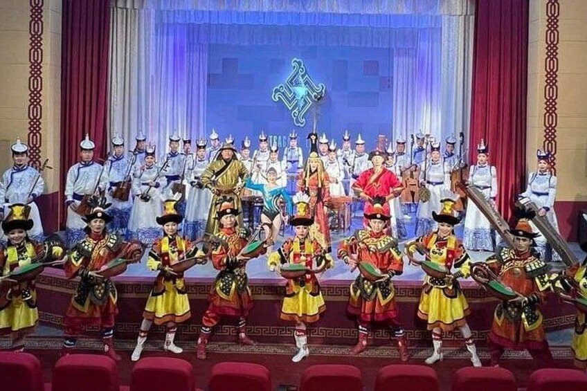 A group of performers in traditional costumes on a stage with a backdrop of curtains and a blue illuminated sign. 5