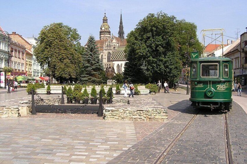 A green tram, a historic building, and a paved square with trees and benches. 5