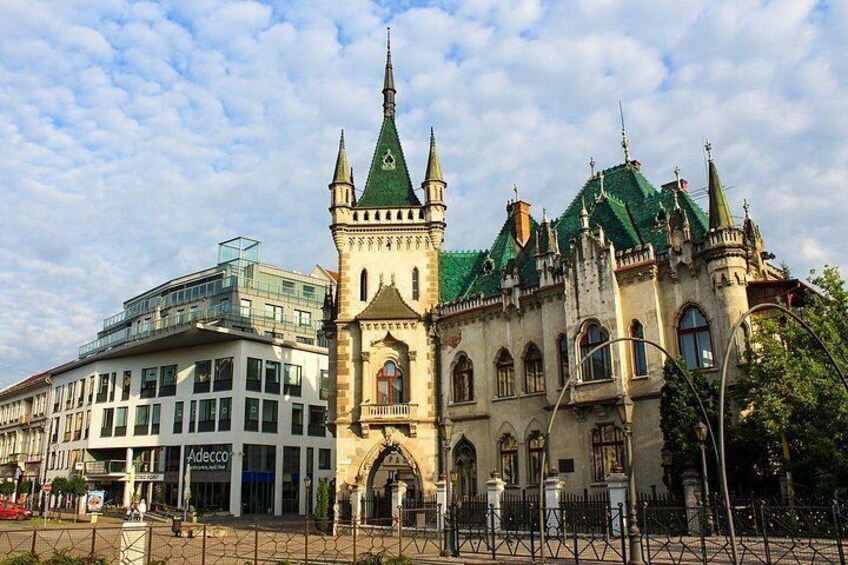 A historic building with a green roof and two towers, a modern glass building, and a fenced area with street lamps. 3
