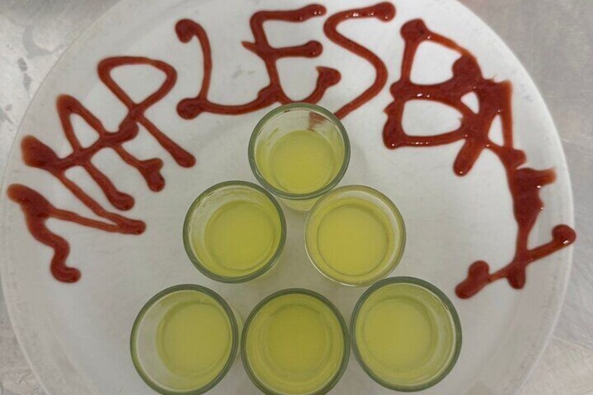 A white plate with five shot glasses of yellow liquid and red text spelling "NAPLES" in ketchup. 5